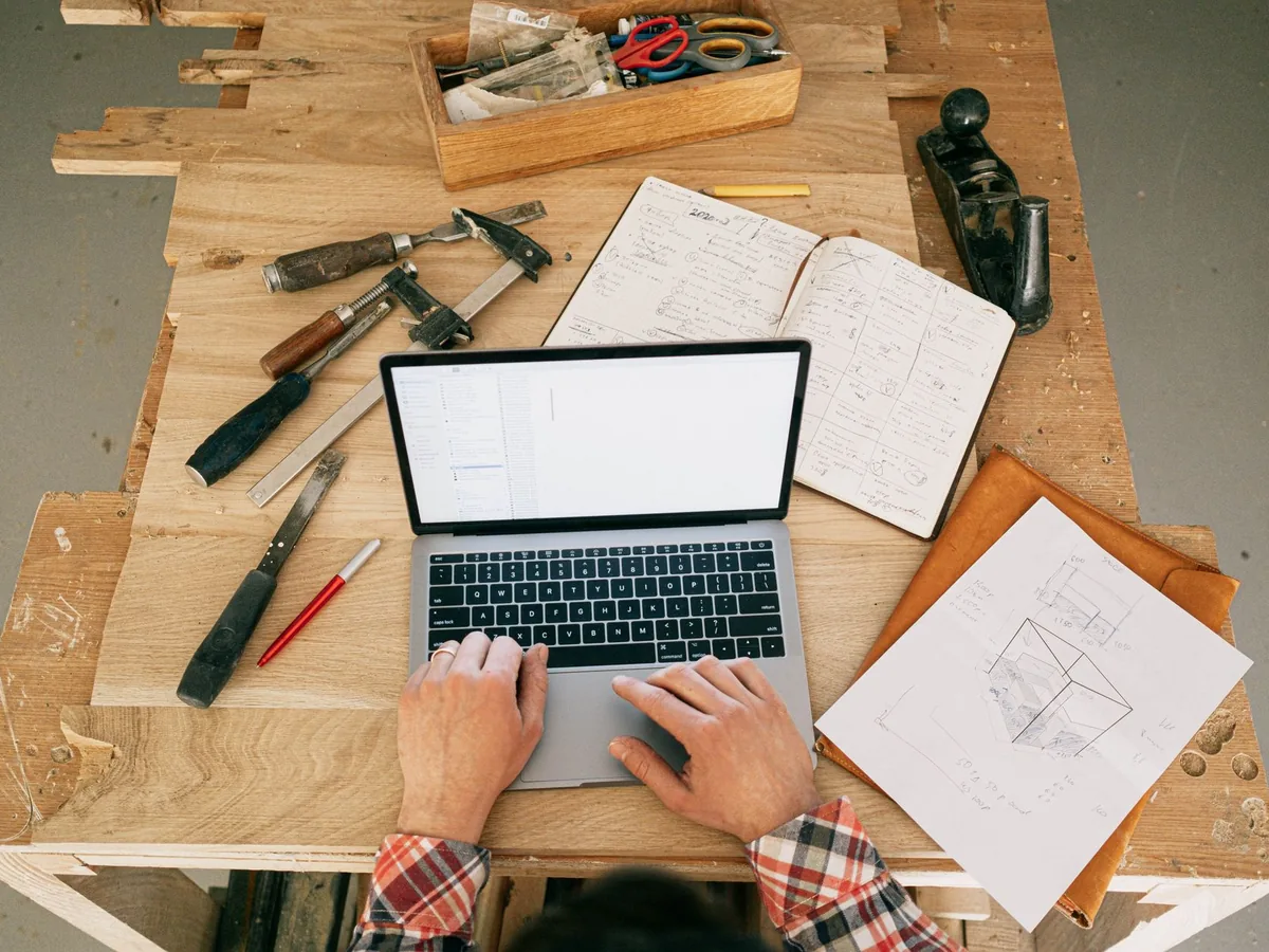 Un menuisier dans son atelier, comparant les plans d'un ouvrage en bois sur un établi avec un devis posé à côté.