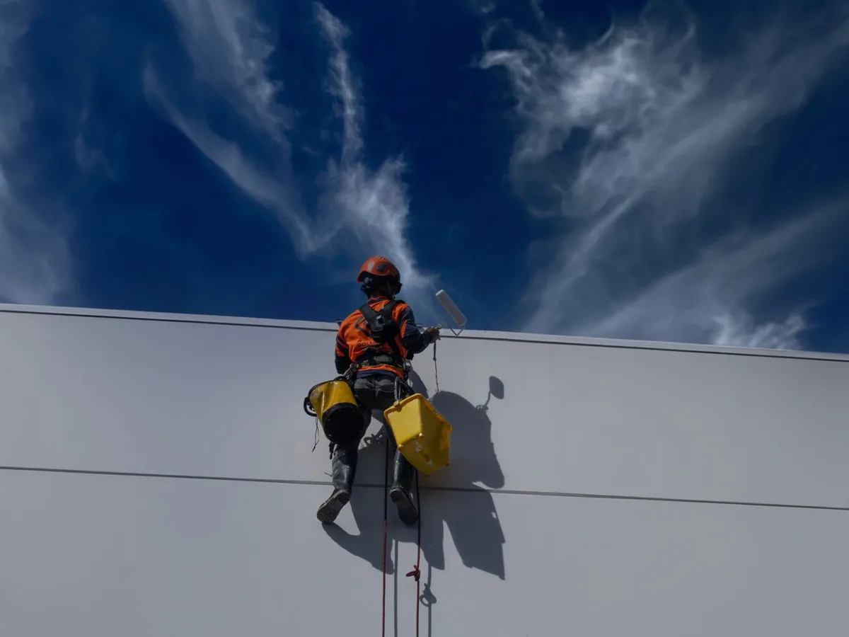 Un peintre en bâtiment établit un devis sur tablette devant une façade à repeindre, avec un chatier=tier en arrière-plan.